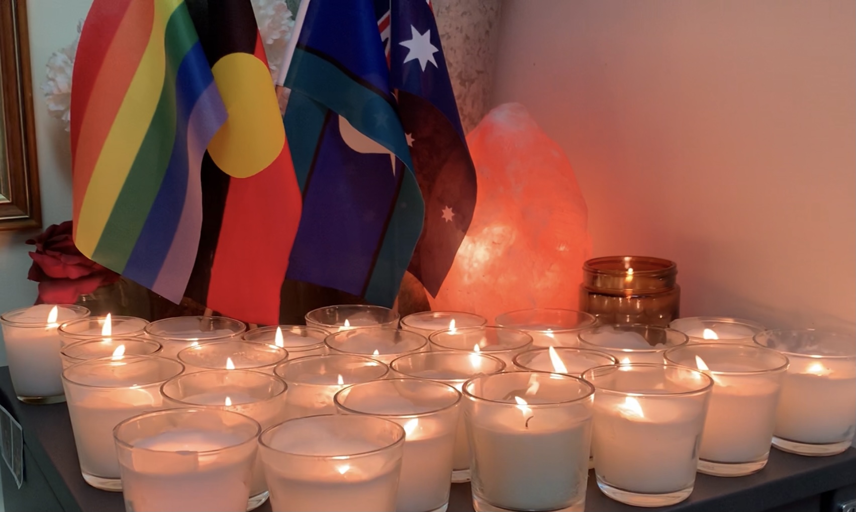 25 votive candles burning with the LGBTIQ, Aboriginal, Torres Strait Island and Australian flags hanging behind them.